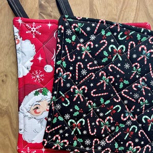 May include: Two quilted Christmas pot holders. One is red with a Santa Claus design and white snowflakes. The other is black with candy canes, green holly, and white snowflakes. Both have black hanging loops.