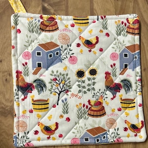 May include: Quilted white pot holder with a farm-themed pattern. The design includes blue barns, red roosters, yellow chicks, sunflowers, and red apples. A yellow fabric loop is attached to the top corner for hanging.