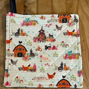May include: Quilted pot holder with a whimsical farm scene. The design features a barn, chickens, cats, pumpkins, and a scarecrow against a striped background. The pot holder has a black loop for hanging and is quilted with a diamond pattern.