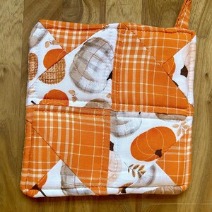 May include: A quilted orange and white pot holder with a pumpkin and leaf design. The pot holder has a loop for hanging.