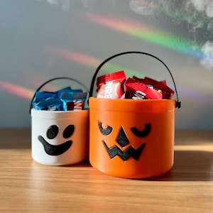 May include: Two Halloween candy buckets. One is white with a black ghost face, and the other is orange with a jack-o'-lantern face. Both have black handles and are filled with wrapped candies.