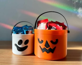 Puede incluir: Dos cubos de Halloween, uno blanco con una cara de fantasma negro y otro naranja con una cara de calabaza. Ambos tienen asas negras y están llenos de caramelos envueltos.