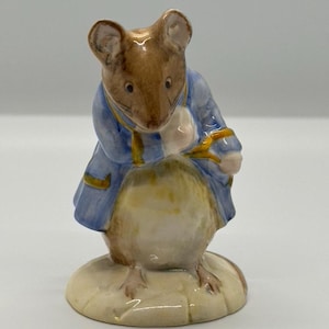 May include: A ceramic figurine of a mouse wearing a blue jacket with yellow trim. The mouse has brown fur and is standing on a cream-colored base. The figurine is a collectible item.
