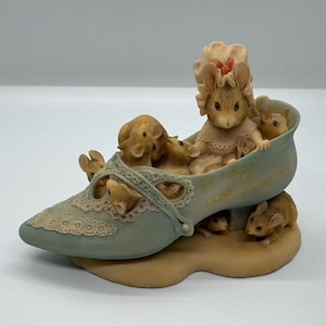 May include: A whimsical figurine of a shoe filled with mice. The shoe is a soft blue-green color with lace detailing. Several mice are inside the shoe, and a few are peeking out. The base of the shoe is a tan color.