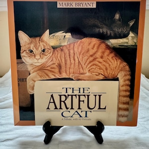 以下が含まれることがあります： マーク・ブライアント著「The Artful Cat: A Tribute, with 60 Portraits」というタイトルの本。表紙には、オレンジ色の虎柄の猫が横たわっている絵が描かれており、背景には黒い猫が眠っています。