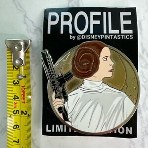 May include: A silver and gold enamel pin featuring Princess Leia from Star Wars. She is holding a blaster and wearing her signature white outfit. The pin is a limited edition collectible.