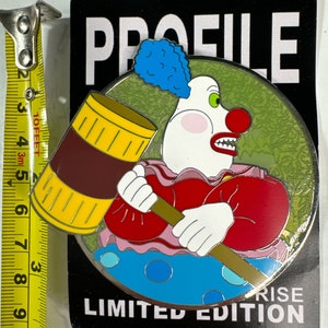 Op de afbeelding: Een ronde, metalen pin met een cartoonclown met een rode neus, groene ogen en een blauwe haardos. De clown houdt een grote, gele en bruine hamer vast. De pin heeft een groene achtergrond met een zwarte rand en de tekst "PROFILE" en "LIMITED EDITION" in wit. De tekst "RISE" is ook zichtbaar in wit.
