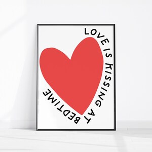 May include: A framed art print featuring a large red heart on a white background. The words "LOVE IS KISSING AT BEDTIME" are written in black, curving around the heart. The frame is black.