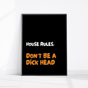 May include: Black framed art print with the text "HOUSE RULES: DON'T BE A D*CK HEAD" in white and orange lettering. The artwork is displayed against a white wall.