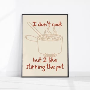May include: A framed print with a beige background and a black frame. The print features a line drawing of a pot with steam and a spoon, with the text "I don't cook but I like stirring the pot" in red.