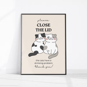 Op de afbeelding: Een ingelijste print met twee cartoonkatten en de tekst "Please Close The Lid". Eén kat is zwart-wit, de andere is grijs en wit. De tekst hieronder luidt "the cats have a drinking problem thank you!".