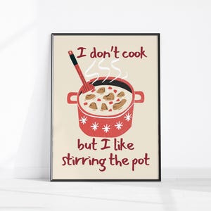 May include: A framed art print featuring a red pot with a wooden spoon stirring food. The text reads "I don't cook but I like stirring the pot" in a playful font. The pot is decorated with white snowflakes.
