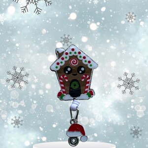May include: A gingerbread house-shaped retractable badge holder with a red Santa hat charm. The gingerbread house has candy cane accents, green and red sprinkles, and a smiling face.