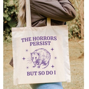 May include: A white tote bag with a purple graphic of an opossum and the text "The Horrors Persist But So Do I".
