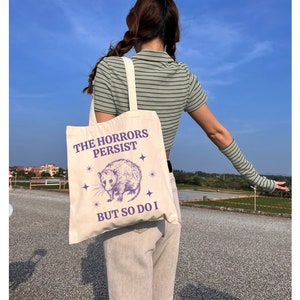 May include: A white canvas tote bag with a purple graphic of a possum and the text "The Horrors Persist But So Do I".