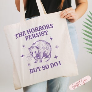May include: A white tote bag with a purple graphic of an opossum and the text "The Horrors Persist But So Do I".