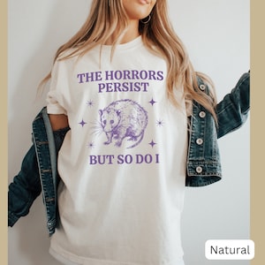 May include: A white t-shirt with a purple opossum graphic and the text "The Horrors Persist But So Do I".