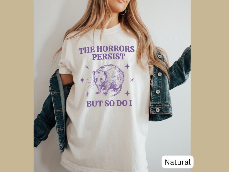 May include: A white t-shirt with a purple opossum graphic and the text "The Horrors Persist But So Do I".