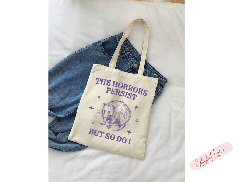 May include: A white tote bag with a purple graphic of a possum and the text "The Horrors Persist But So Do I".
