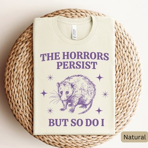 May include: A light beige t-shirt with a purple graphic of an opossum and the text "The Horrors Persist But So Do I".