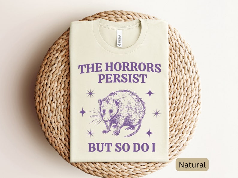 May include: A light beige t-shirt with a purple graphic of an opossum and the text "The Horrors Persist But So Do I".