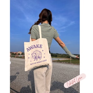 K&ouml;nnte beinhalten: Eine wei&szlig;e Canvas-Tragetasche mit einer lilafarbenen Grafik eines Opossums und dem Text "Awake but at what cost".