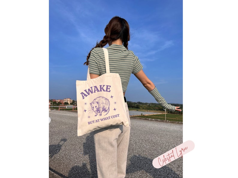 K&ouml;nnte beinhalten: Eine wei&szlig;e Canvas-Tragetasche mit einer lilafarbenen Grafik eines Opossums und dem Text "Awake but at what cost".