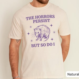 May include: A white t-shirt with a purple opossum graphic and the text "The Horrors Persist But So Do I".