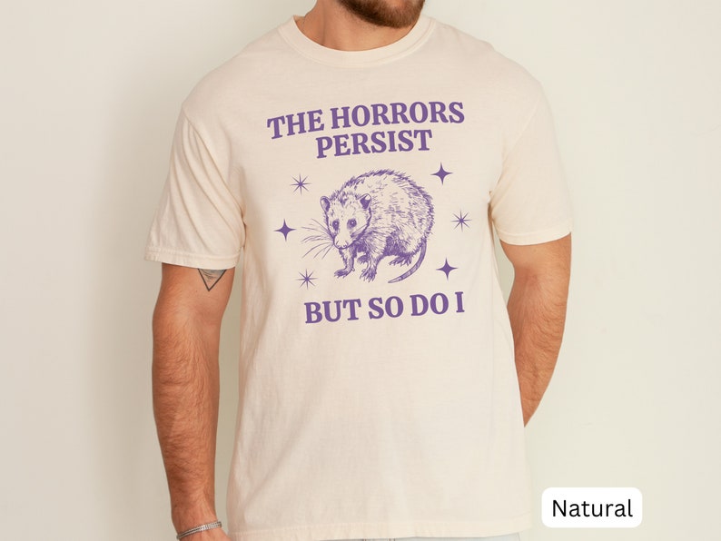 May include: A white t-shirt with a purple opossum graphic and the text "The Horrors Persist But So Do I".