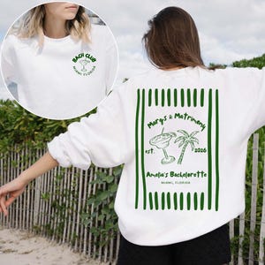May include: White sweatshirt with green text and graphics. The front features a small "Bach Club" logo. The back has a larger design with a cocktail, palm tree, and the text "Margs & Matrimony" and "Amelia's Bachelorette" in green.