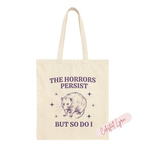 May include: A beige tote bag with a purple graphic of a possum and the text "The Horrors Persist But So Do I".