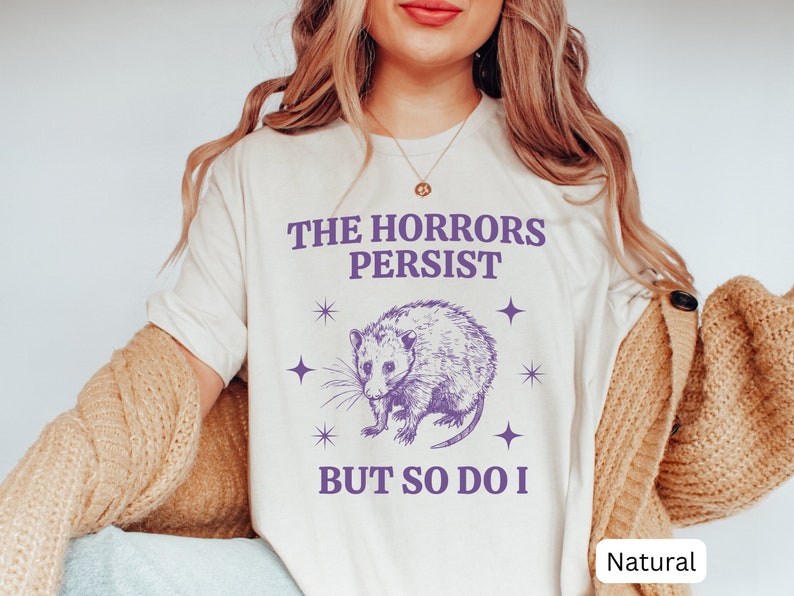 May include: A white t-shirt with a purple graphic of an opossum and the text "The Horrors Persist But So Do I".