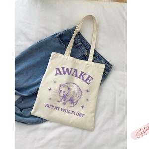 K&ouml;nnte beinhalten: Eine wei&szlig;e Einkaufstasche mit einer lila Grafik eines Opossums und dem Text "Awake but at what cost".