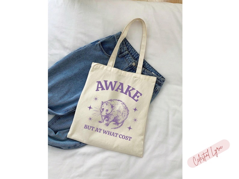 K&ouml;nnte beinhalten: Eine wei&szlig;e Einkaufstasche mit einer lila Grafik eines Opossums und dem Text "Awake but at what cost".