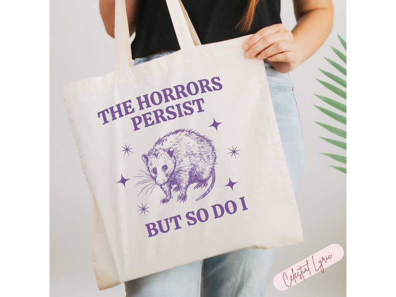 May include: A white tote bag with a purple graphic of an opossum and the text "The Horrors Persist But So Do I".