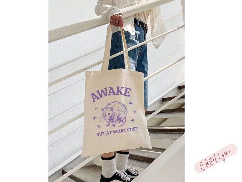 K&ouml;nnte beinhalten: Eine wei&szlig;e Einkaufstasche mit einer lila Grafik eines Opossums mit Sternen und dem Text "Awake but at what cost".