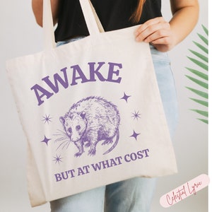 K&ouml;nnte beinhalten: Eine wei&szlig;e Einkaufstasche mit einer lila Opossum-Illustration und dem Text "AWAKE BUT AT WHAT COST" in lila Schrift. Die Tasche hat ein lila Sternendesign.