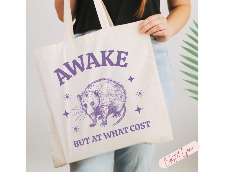 K&ouml;nnte beinhalten: Eine wei&szlig;e Einkaufstasche mit einer lila Opossum-Illustration und dem Text "AWAKE BUT AT WHAT COST" in lila Schrift. Die Tasche hat ein lila Sternendesign.