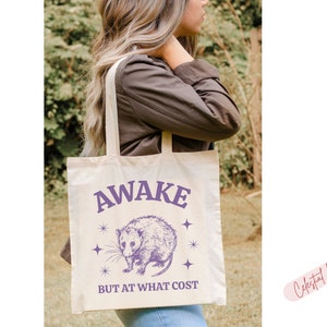 K&ouml;nnte beinhalten: Eine wei&szlig;e Canvas-Tragetasche mit einer lilafarbenen Grafik eines Opossums und dem Text "Awake but at what cost".
