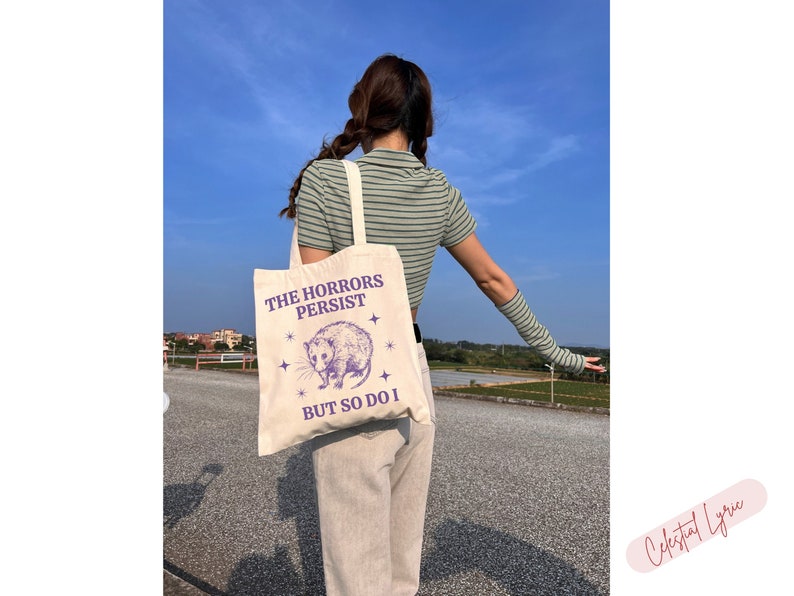 May include: A white canvas tote bag with a purple graphic of a possum and the text "The Horrors Persist But So Do I".