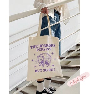May include: A white canvas tote bag with a purple graphic of a possum and the text "The Horrors Persist But So Do I" with stars around the graphic.