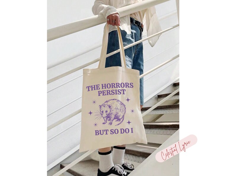 May include: A white canvas tote bag with a purple graphic of a possum and the text "The Horrors Persist But So Do I" with stars around the graphic.