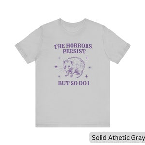 May include: A light gray t-shirt with a purple opossum graphic and the text "The Horrors Persist But So Do I".