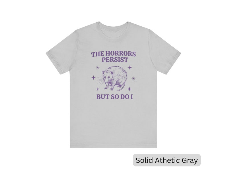 May include: A light gray t-shirt with a purple opossum graphic and the text "The Horrors Persist But So Do I".