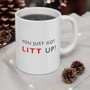 Può includere: Tazza da caffè in ceramica bianca con manico nero. La tazza ha la scritta "You just got LITT UP!" stampata in nero e rosso.