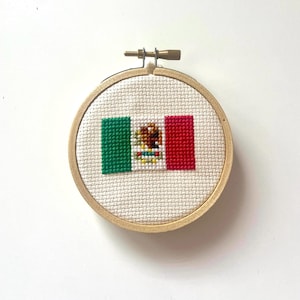 May include: A small, round wooden hoop with a white fabric stretched across it. The fabric has a cross-stitch design of the Mexican flag, featuring green, white, and red stripes with the Mexican coat of arms in the center.