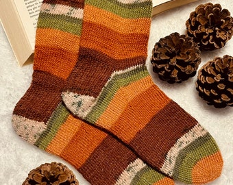 May include: A pair of hand-knitted socks with a striped pattern in shades of brown, orange, green, and white. The socks are displayed with pine cones and an open book, suggesting a cozy, autumnal theme.