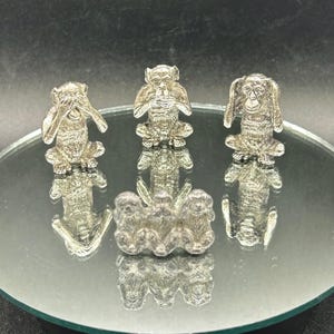 May include: Silver figurines of the three wise monkeys: see no evil, hear no evil, speak no evil. The monkeys are in a seated position with their hands covering their eyes, ears, and mouth. The figurines are displayed on a reflective surface.