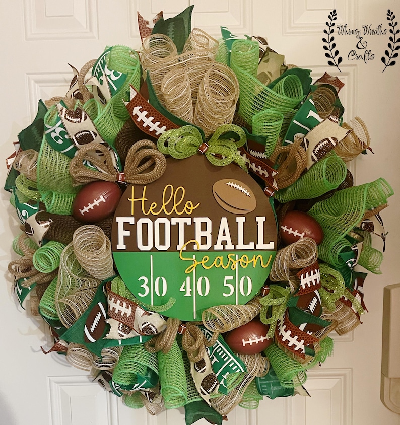 May include: A decorative football-themed wreath with green, brown, and tan mesh. The wreath features miniature footballs, ribbons, and a central sign that reads "Hello FOOTBALL Season" with yard markers. A festive decoration for the football season.