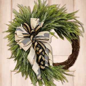 May include: A decorative wreath featuring a circular grapevine base, lush green foliage, and a large bow. The bow is made of beige and black patterned ribbons, adding a touch of elegance to the natural elements. This wreath is suitable for seasonal home decor.